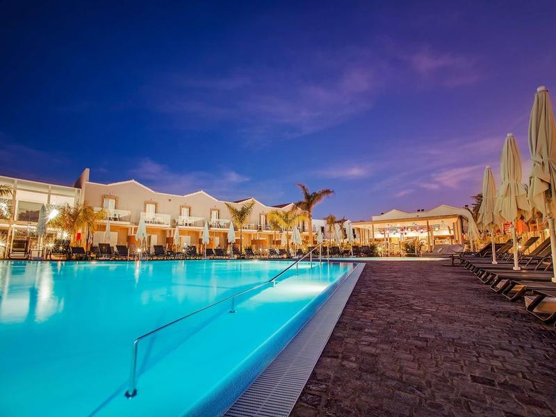 Wieczorny widok oświetlonego basenu hotelowego z leżakami i budynkami w tle.