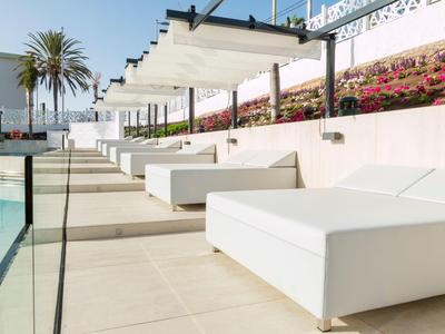 Chaises longues blanches au bord de la piscine sous un auvent avec des palmiers et des fleurs en arrière-plan.