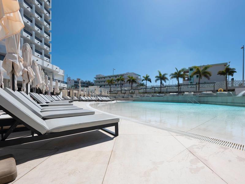 Espace piscine d'hôtel ensoleillé avec chaises longues et palmiers sous un ciel bleu clair.