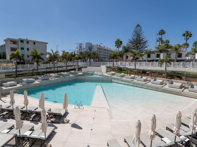 Grande piscine extérieure avec chaises longues et parasols dans un hôtel sous un ciel bleu clair.