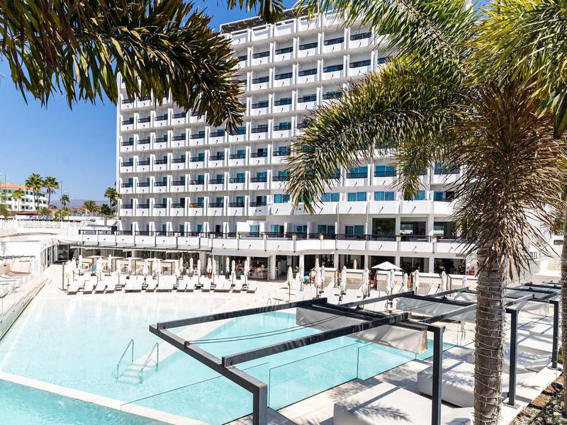 Bâtiment hôtelier moderne avec grande piscine extérieure et palmiers par temps ensoleillé.