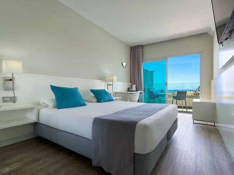 Chambre d'hôtel moderne avec lit double, coussins bleus et vue sur la mer depuis le balcon.