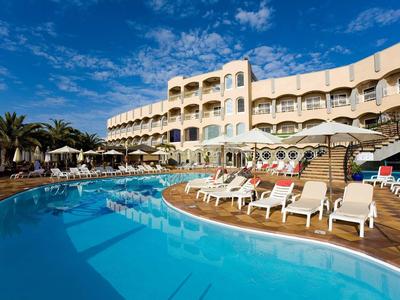 Hôtel avec grande piscine, chaises longues et palmiers sous un ciel bleu.