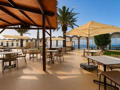 Buitenterras met tafels, stoelen, parasols en zeezicht onder een heldere lucht.