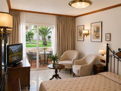 Lichte hotelkamer met tweepersoonsbed, twee fauteuils, tafel, tv en balkon met uitzicht op tuin.