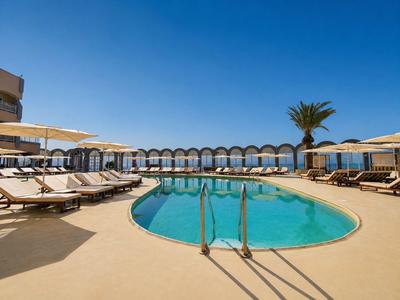 Rond hotelzwembad met ligstoelen en parasols onder een heldere blauwe lucht.