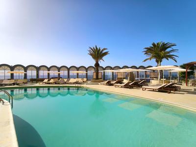Ronde hotelzwembad met turquoise water, ligstoelen en palmbomen onder een heldere lucht.