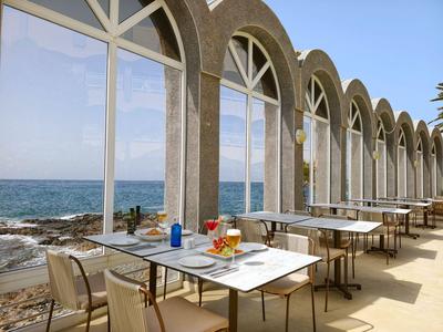 Hotelterras met tafels en stoelen aan zee onder een heldere hemel.