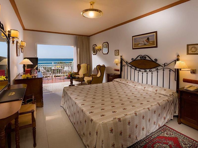Bright hotel room with double bed, seating area, and sea view from the balcony.