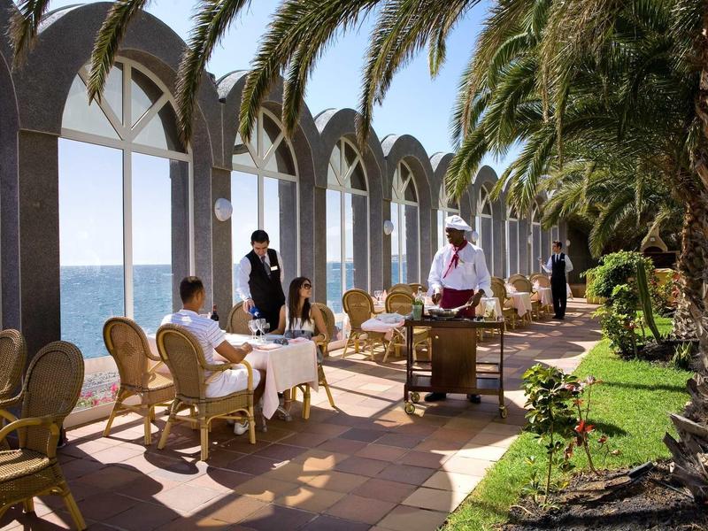 Host serves food in a shaded outdoor hotel area with sea view and palm trees.