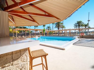 Moderna zona de piscina exterior con tumbonas y decoración de palmeras bajo sombrillas.