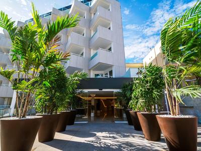 Entrada moderna de hotel con grandes plantas en macetas y edificio blanco bajo cielo azul.