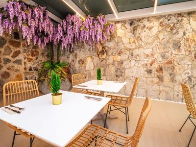 Jadalnia z białymi stołami, bambusowymi krzesłami i wiszącymi fioletowymi kwiatami przy kamiennej ścianie.