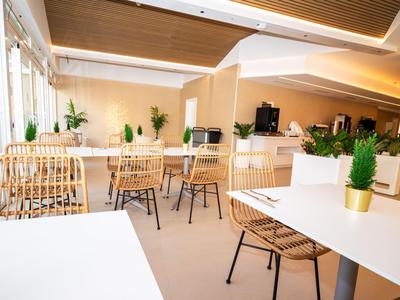 Nowoczesna jadalnia z białymi stołami, krzesłami z rattanu i zielonymi roślinami w doniczkach.