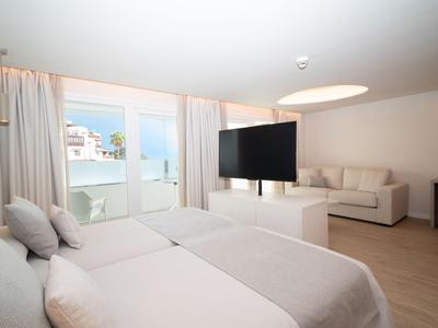 Jasny pokój hotelowy z dwoma łóżkami, telewizorem i sofą z widokiem na balkon i morze.