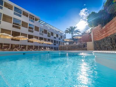 Hotel met blauw buitenzwembad, parasols en palmbomen onder een heldere lucht.