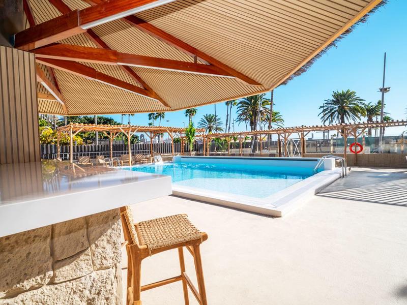 Moderna zona de piscina exterior con tumbonas y decoración de palmeras bajo sombrillas.