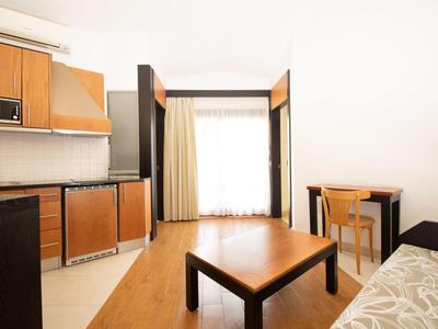 Lichte hotelkamer met kitchenette, eettafel, bank en groot raam met gordijnen.