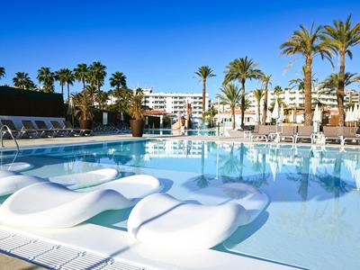 Ein großer Pool mit weißen Liegen und Palmen vor hellen Hotelgebäuden unter klarem blauem Himmel.