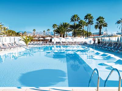 Gran piscina de hotel con palmeras y tumbonas bajo un cielo azul claro.