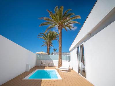 Moderner Hotelbereich mit kleinem Pool, Palmen und weiß gestrichenen Wänden unter klarem blauem Himmel.
