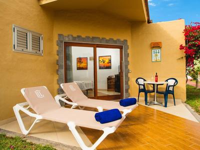 Twee ligstoelen met blauwe handdoeken en een kleine tafel met twee stoelen voor een geel huis.