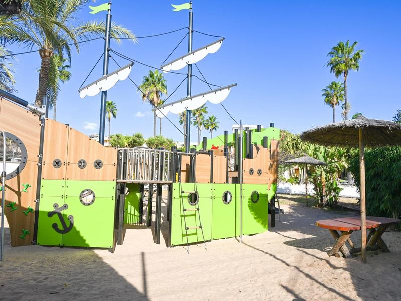 Kleurrijke speeltuin voor kinderen in de vorm van een schip met palmbomen en parasols op de achtergrond.