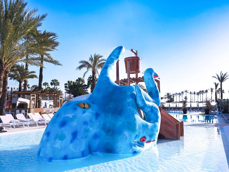Kleurrijke speelzone met blauwe walvisglijbaan en palmbomen bij het hotelzwembad.