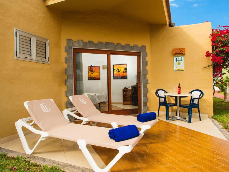 Twee ligstoelen met blauwe handdoeken en een kleine tafel met twee stoelen voor een geel huis.