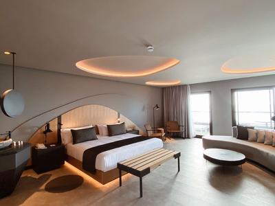 Modernes, helles Hotelzimmer mit großem Bett, Sofalounge, runden Lampen und Fensterfront.