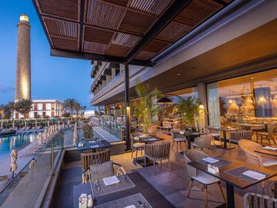 Restaurantterrasse mit Holzdecke, Tischen, Stühlen und Blick auf beleuchteten Leuchtturm bei Dämmerung.