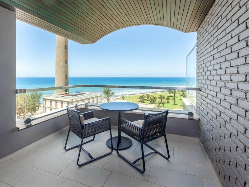 Balcón con mesa y dos sillas con vista a la playa y el mar bajo cielo azul.