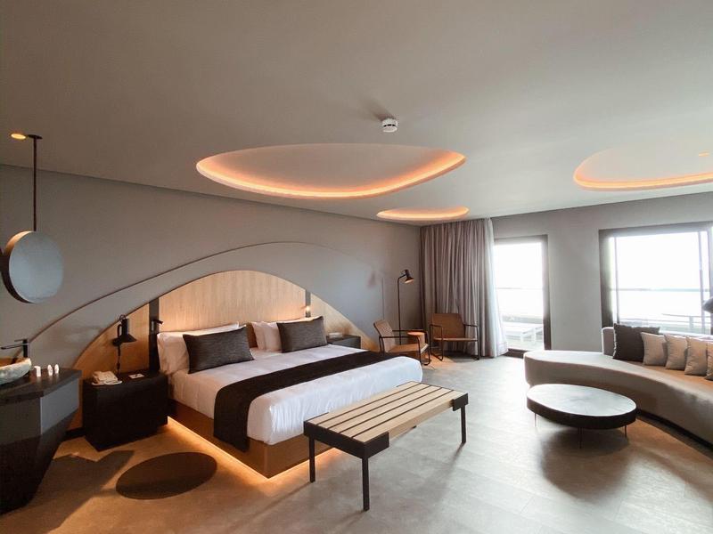 Habitación de hotel moderna con cama grande, sofá en esquina y luz cálida en el techo.