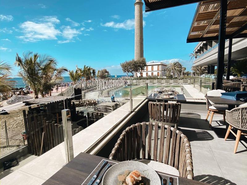 Terraza de un restaurante con mesa, sillas y vista a un faro en un día soleado.
