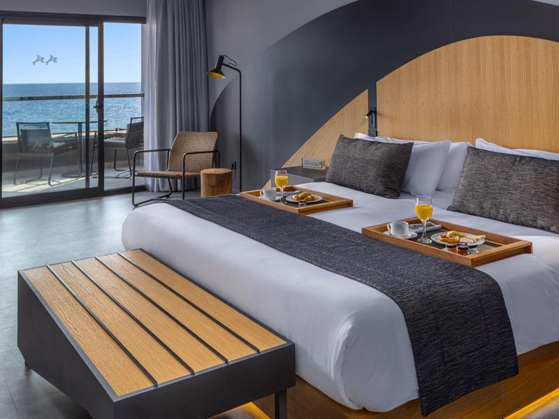 Habitación de hotel moderna con vista al mar, cama de diseño, muebles de madera y bandejas de desayuno.