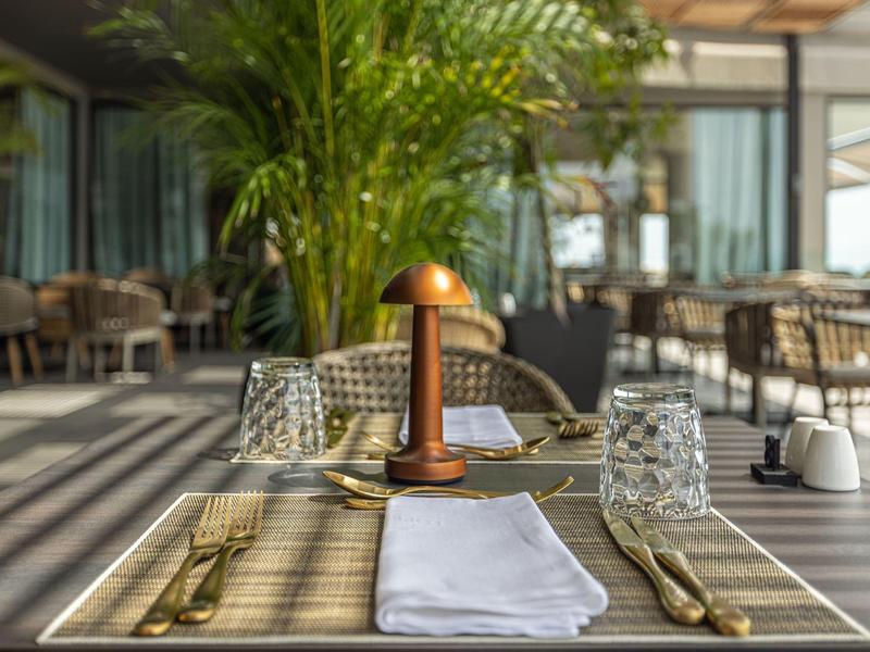 Mesa preparada con manteles y cubiertos en un restaurante al aire libre con plantas y sillas.