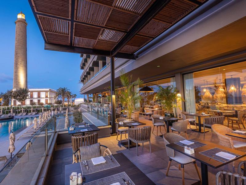 Terraza del hotel por la tarde con mesas y vista a una torre iluminada junto al water.