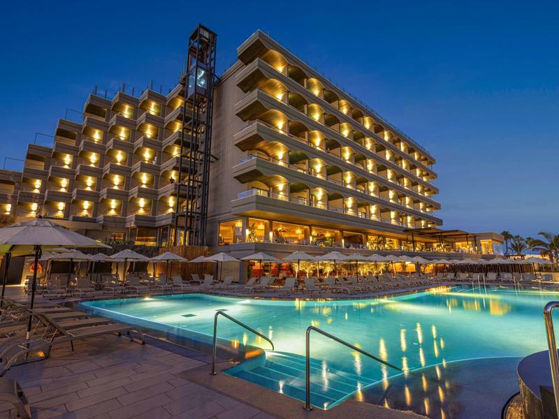 Hotel con luces encendidas al atardecer junto a una piscina iluminada y sombrillas junto a las tumbonas.