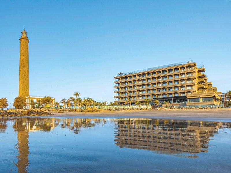 Edificio de hotel y faro reflejados claramente en agua tranquila bajo cielo azul.