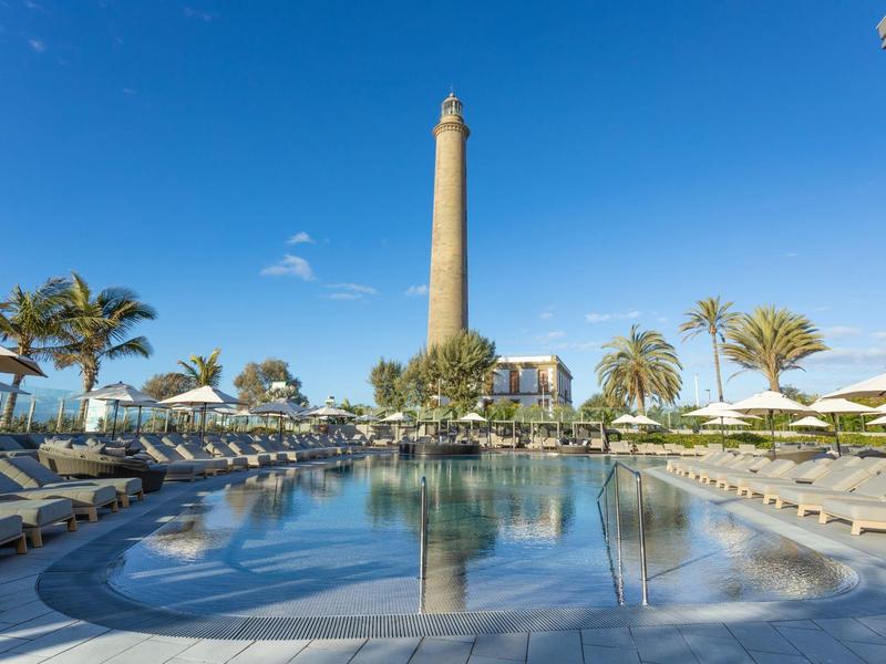 Gran piscina con tumbonas y palmeras, alto faro al fondo bajo cielo despejado.