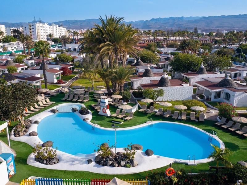 Resort with kidney-shaped pool, palm trees, sun loungers, and nearby low-rise buildings.