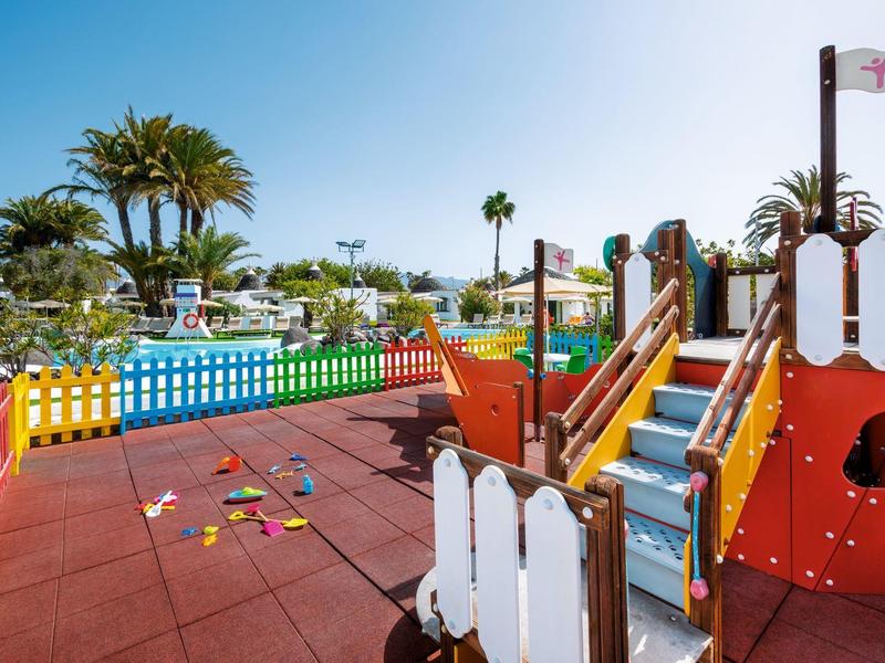 Colorful playground with climbing structure near a pool and palm trees under a clear sky.