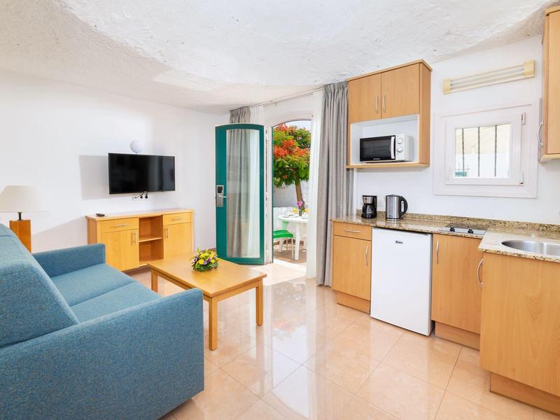 Cozy living room with blue sofa, wooden furniture, kitchenette, and open door to outdoor area.