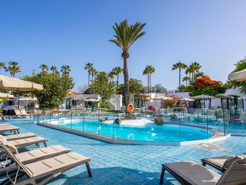 Outdoor pool area with sun loungers, umbrellas, and palm trees under a clear blue sky.