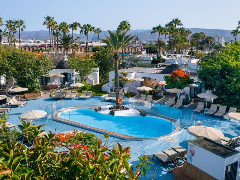 Resort with a large pool, sunbeds, palm trees, and surrounding greenery under clear skies.