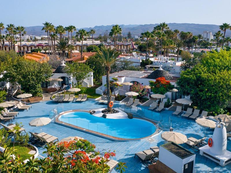 Outdoor hotel pool surrounded by lounge chairs, umbrellas, palm trees, and lush greenery under clear skies.