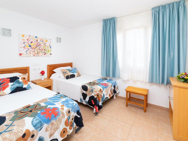 Bright hotel room with two single beds, colorful bedspreads, blue curtains, and wooden furniture.