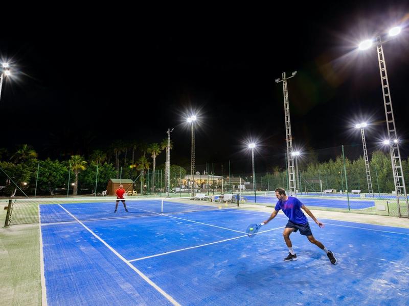 Two people playing tennis on a blue court under bright night lights with palm trees in the background