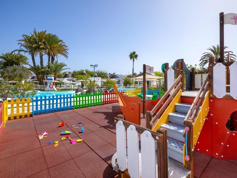 Children's pool area with pirate ship slide, colorful fence, and palm trees under clear sky.
