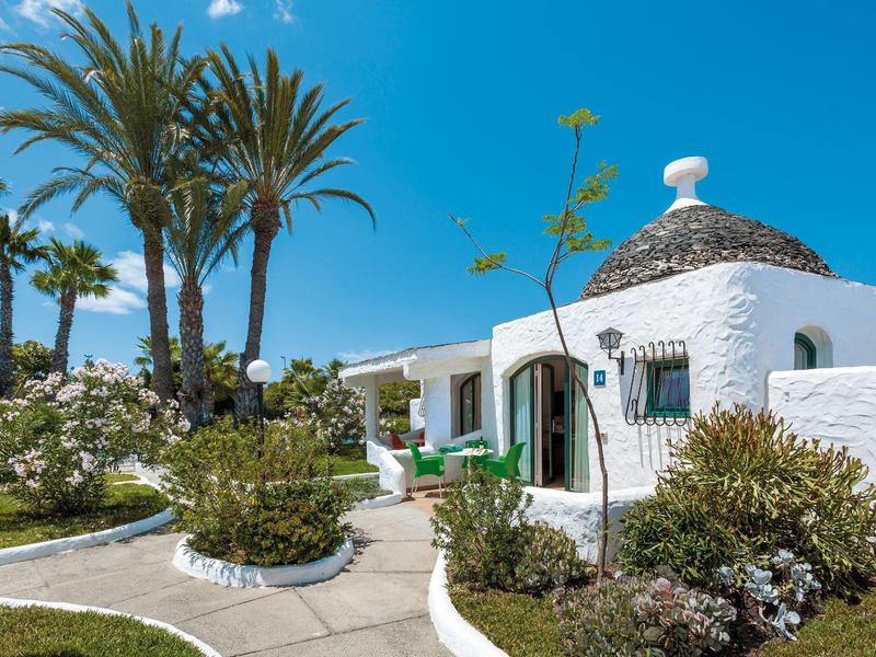 White dome-shaped houses with thatched roofs surrounded by palm trees and garden paths.